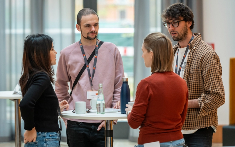 Four people stand around a high table at a networking event, talking and drinking coffee.